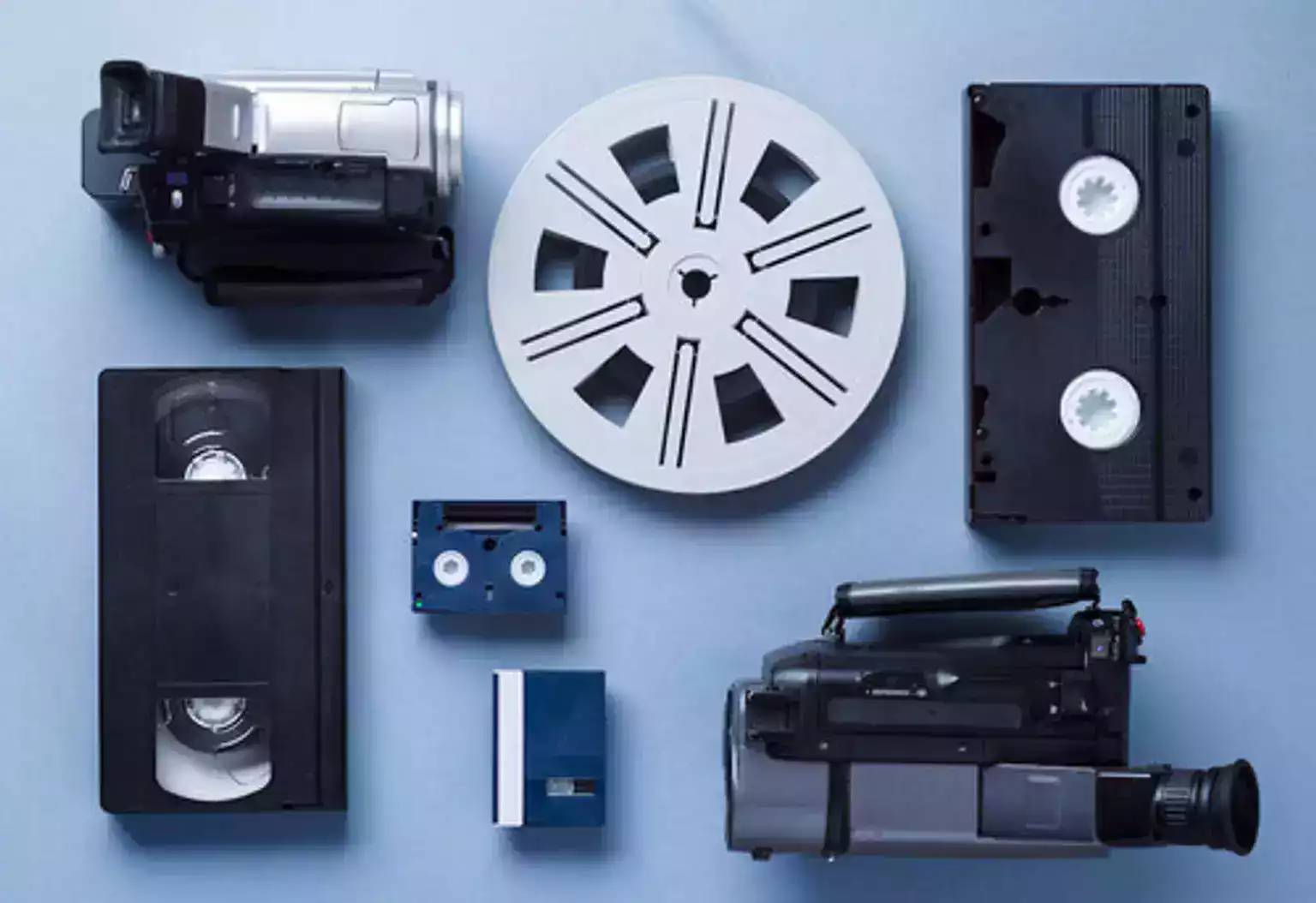 Various vintage video recording equipment and media, including camcorders, VHS tapes, a reel, and a cassette, arranged on a blue surface.